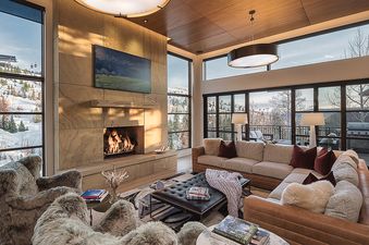 A spacious, luxurious living room features a stone fireplace, large windows overlooking a snowy mountain landscape, and comfortable seating arrangements, perfect for relaxation.