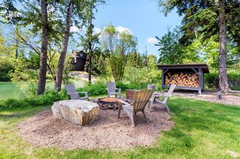 A serene outdoor fire pit area at Landing Unit A with Adirondack chairs, a stocked woodshed, and a tranquil body of water bordered by lush trees.