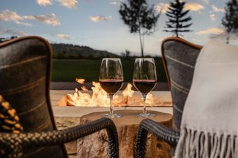 Two chairs face a glowing fire pit with two glasses of red wine, set against a backdrop of hills and a sunset sky.