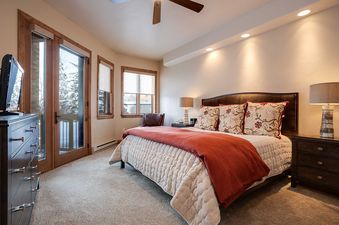 A cozy bedroom with a king-sized bed, two nightstands with lamps, a dresser with a TV, and a glass door leading to a balcony with a winter view.
