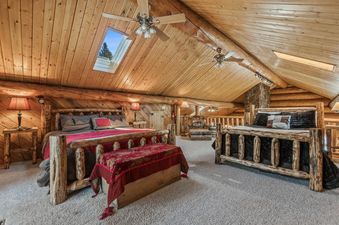The primary bedroom at Stillwater Log Home features two log-framed beds, log furniture, and a skylight on a vaulted wooden ceiling, all contributing to a rustic aesthetic.