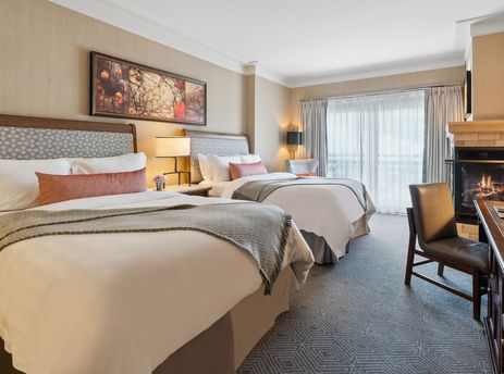 A luxurious hotel room features two queen beds with crisp white linens, accent pillows, and throws, a working fireplace, a desk with a chair, and a view of the mountains through the balcony.