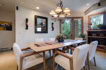A dining room at Chateau Chamonix CX311 with a wooden table, cream chairs, chandelier, and large windows looking out to trees.