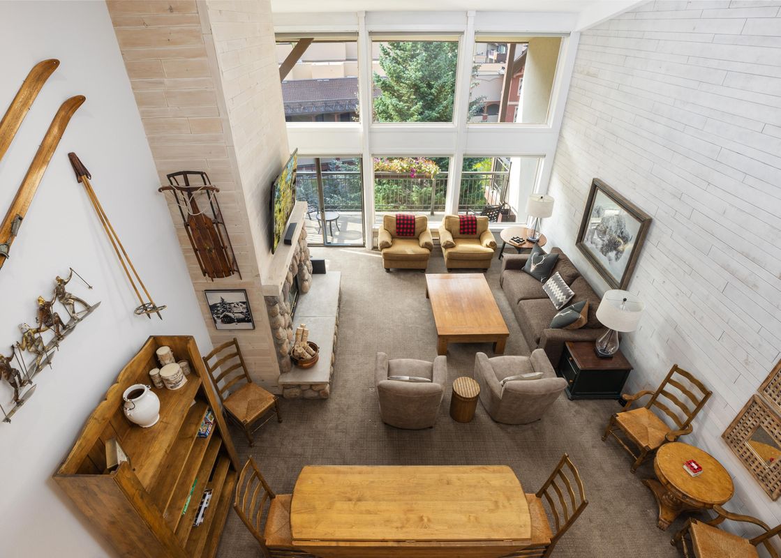 A high-angle view of the Montaneros 412 living room, featuring a fireplace, a brown sectional sofa, several armchairs, wooden tables, and large windows with exterior views.