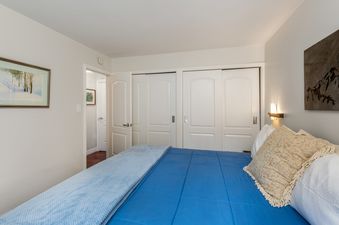 A comfortable blue bed with decorative pillows and a light blue throw blanket, flanked by white closet doors and a painting on the wall.