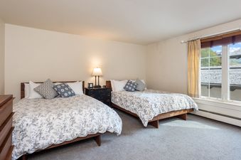 The image shows a bedroom featuring two comfortable beds, each adorned with white and blue floral bedding and decorative pillows, bathed in natural light from a large window overlooking a lush outdoor landscape.