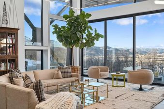 A luxurious living room features plush, light-colored sofas, tiered glass coffee tables, and two swivel armchairs, all set against a backdrop of floor-to-ceiling windows with panoramic mountain views.
