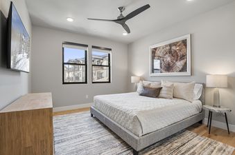 A modern and serene bedroom features a queen-sized bed with white bedding, a unique marble-topped bedside table with a lamp, and a large abstract tree ring art piece on the wall, with windows providing natural light.