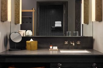 A stylish bathroom vanity features a large mirror, dark countertop, white sink, and illuminated wall sconces, with a wooden stool tucked underneath.