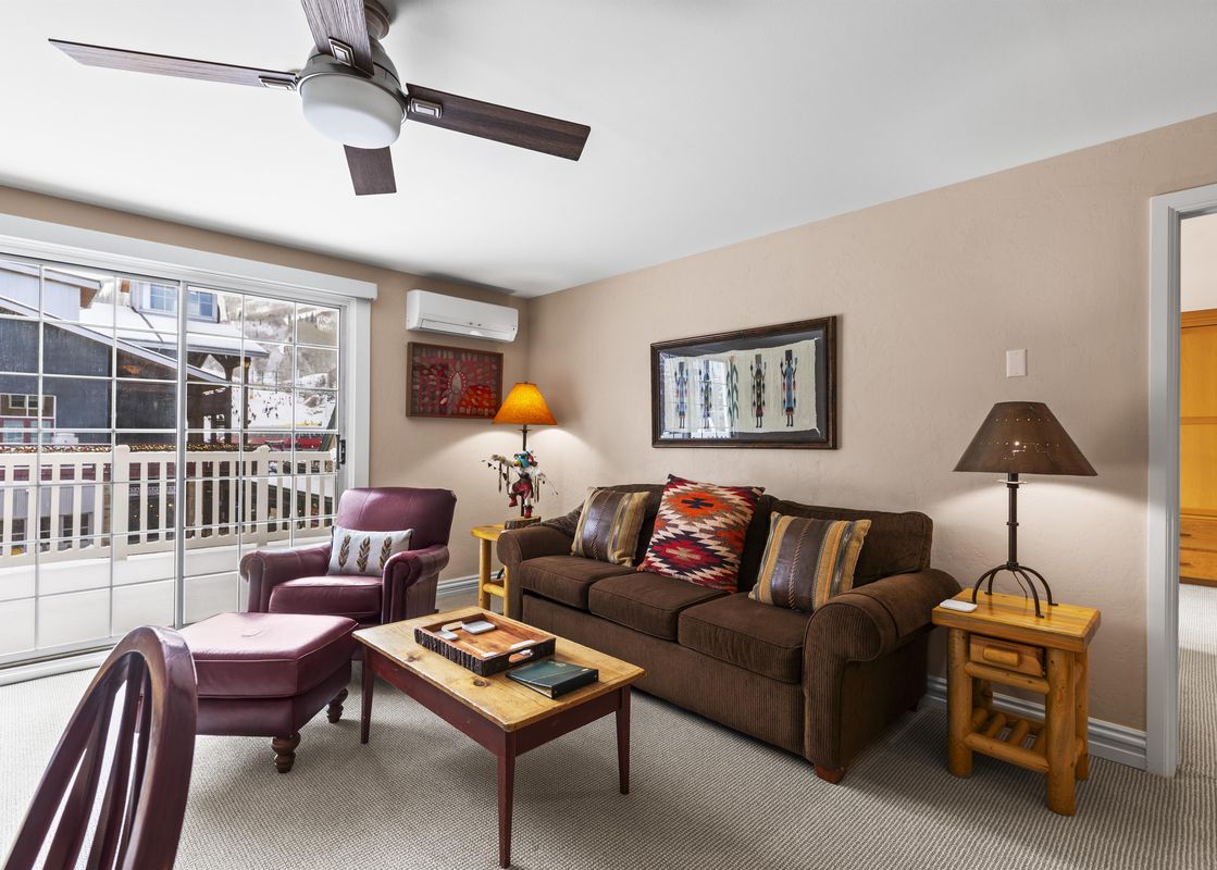 The living area of Resort Plaza 5051 featuring a brown sofa, burgundy armchair, rustic wooden coffee table, and sliding glass doors with a view of a snowy landscape.