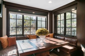 A cozy dining nook with a large wooden table and brown leather banquette seating, featuring big windows that offer a view of the outdoors.