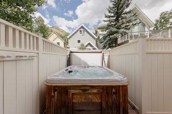 An outdoor hot tub with bubbling water is nestled within a white privacy fence at Smugglers Hideaway, surrounded by houses and mountains under a blue sky.