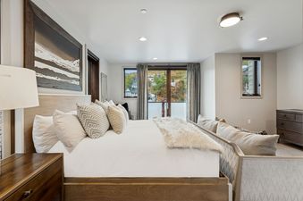 A king-sized bed with white bedding and numerous pillows sits in a bedroom with a view of a mountainous landscape and houses from a window and glass door leading to a balcony.