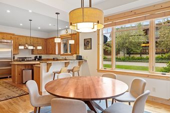 A modern kitchen with wooden cabinetry, stainless steel appliances, a breakfast bar, and a dining area with a round wooden table and four chairs, all brightly lit by natural light from large windows.