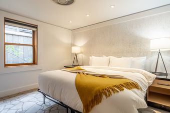 A modern bedroom with a king-size bed, white linens, a yellow throw, and two bedside lamps. A window offers a view of outdoor greenery, and the room features recessed lighting and textured wallpaper.