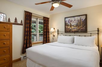 A cozy bedroom in Montaneros 308 featuring a large bed with white linens, a brass headboard, a window with patterned curtains revealing green trees, and a mountain landscape painting.