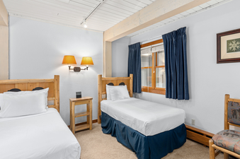 A twin bedroom at Top of the Village Leaf 401 with two beds, a nightstand, lamps, and a window with blue curtains.