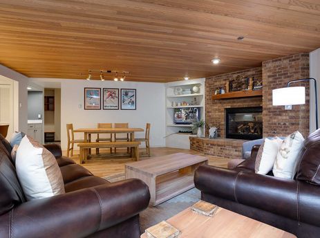 A comfortable living room with brown leather couches, a brick fireplace, and a wooden dining table, all under a warm wood-paneled ceiling.