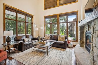 The living room of Whitefish Ski Haus features two brown leather sofas, a glass-top coffee table, a stone fireplace, and large windows with wooden frames overlooking a wooded area.