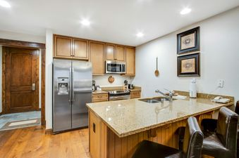A modern kitchen at BlueSky Breckenridge 410 showcasing stainless steel appliances, granite countertops, and light wood cabinetry, with an island featuring a double sink and bar stools.