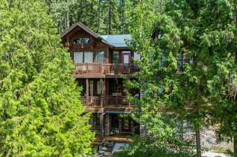A multi-level wooden cabin, Cottonwood 204, is nestled among lush green trees, featuring several balconies and a dark green roof.