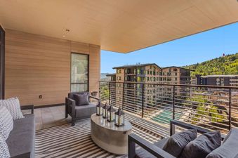 A modern outdoor balcony at One Snowmass East 704 features comfortable seating, a stylish concrete coffee table with lanterns, and a striped rug, all overlooking a scenic mountain landscape with buildings and lush greenery under a clear blue sky.