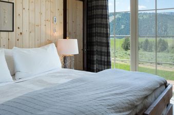 A cozy bedroom in The Shooting Cabin features a comfortable bed, wood-paneled walls with botanical art, and a large window overlooking a scenic mountain landscape.