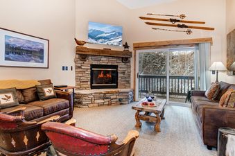 A cozy living room at Snow Flower 156 Park City with a stone fireplace, brown sofas, a rustic coffee table, and a sliding glass door leading to a snowy balcony view.