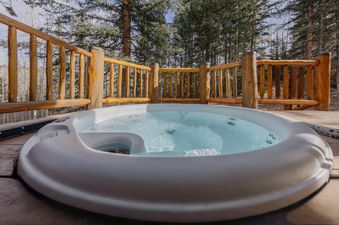 A bubbling hot tub on a wooden deck, surrounded by a rustic log railing and a backdrop of pine trees.