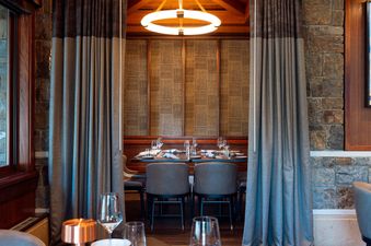 A dining table is set within a cozy, private alcove, flanked by elegant drapes and framed by rustic stone walls and dark wood.