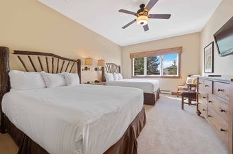 A hotel bedroom features two queen beds with log-style headboards, two wall lamps, a large window with an exterior view, a wooden dresser, a wall-mounted TV, and a ceiling fan.