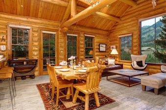 A rustic dining area with a natural log table and wooden chairs, flowing into a cozy living space with a leather sofa and armchair, all within a log cabin with large windows showcasing mountain views.