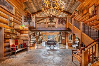 A grand foyer at Elk Mountain Lodge with log walls, a large antler chandelier, a stone floor, and a wooden staircase leading to an upper level decorated with saddles and chaps.