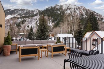 An expansive outdoor terrace featuring a large L-shaped sofa, two armchairs, and a dining table with chairs, set against a backdrop of snow-covered mountains and evergreen trees under a partly cloudy sky.