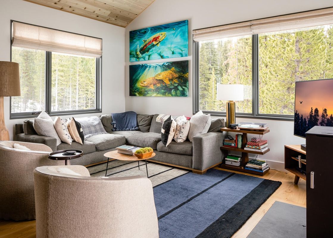 A spacious living area in Moonlight Basin 27 Lake Cabin, featuring a plush sectional sofa with throw pillows, a coffee table with a vase and greenery, and two cozy armchairs, all surrounding a  blue rug. The room boasts large windows that offer breathtaki