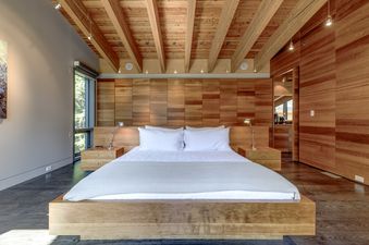 A serene bedroom in Highpoint Chalet features a queen-sized bed with white linens, flanked by wooden nightstands and chrome lamps, set against a wooden accent wall and ceiling with exposed beams, and a large window offering an exterior view.
