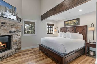 A cozy bedroom with a king-sized bed, stone fireplace with a roaring fire, a TV, and two windows with views of trees.