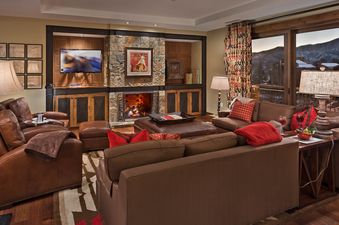 A luxurious and cozy living room featuring brown leather seating, a stone fireplace, and a large window offering a view of snowy mountains.