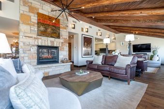 A spacious living room featuring a stone fireplace, brown leather sofas, and an octagonal coffee table, illuminated by a unique chandelier and sconces.