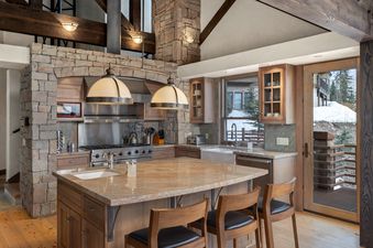 The kitchen at Rocky Road Hideaway features an island with seating, stainless steel appliances, and stone accents, with a large window looking out to a snowy landscape.