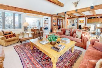 A spacious and elegantly furnished living room in the Trails Edge Vacation Home, featuring plush seating, a large wooden coffee table, exposed wooden beams, and expansive windows offering views of a snowy landscape.