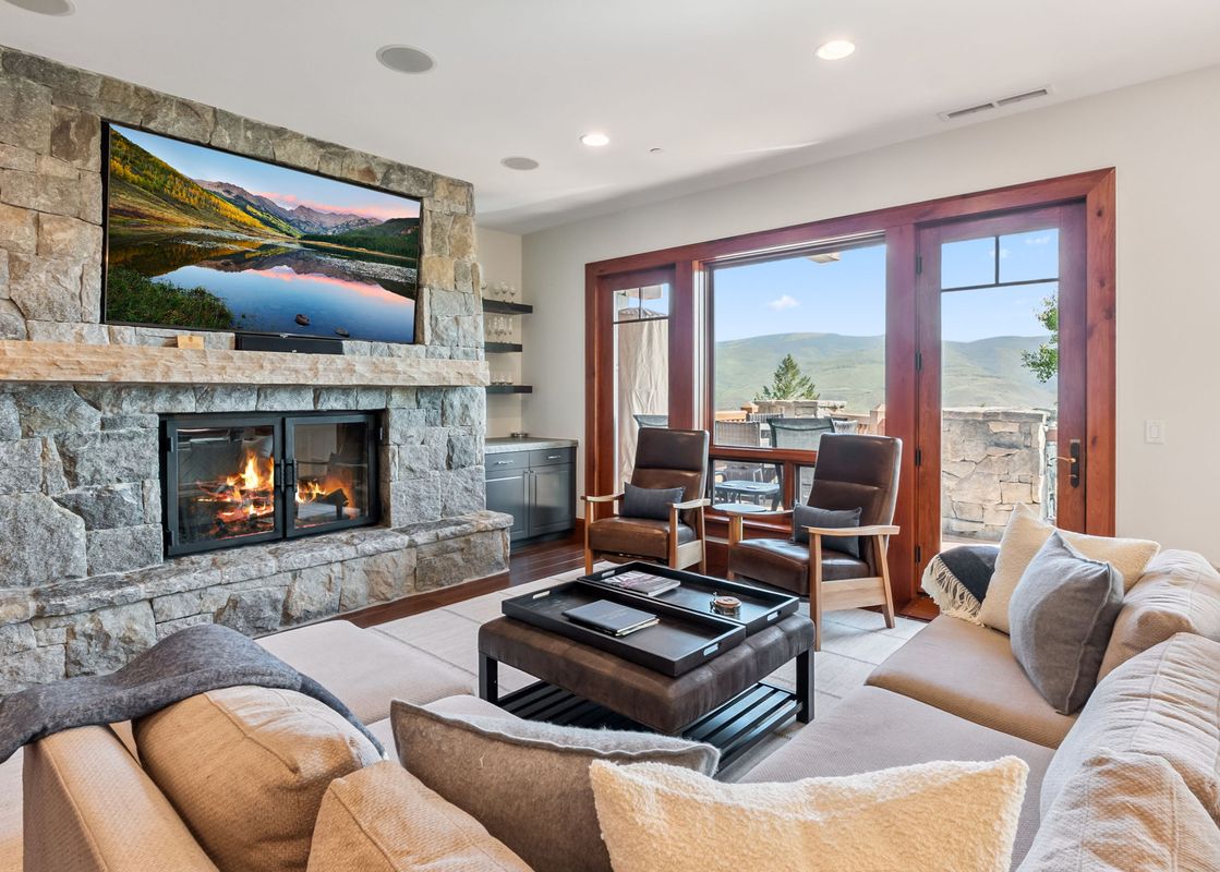 A luxurious living room at Blue Bird Vista at Hummingbird Lodge features a large sectional sofa, a stone fireplace with a TV above, and glass doors opening to a balcony with mountain views.