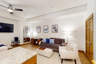 A cozy living area features a gray sofa with blue pillows, a white armchair, and a flat-screen TV mounted on the wall, with ski-themed artwork decorating the walls.