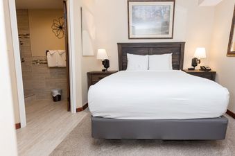 A comfortable bedroom at the Beaver Run Resort Studio features a large bed with white linens and bedside tables with lamps, with an open doorway leading to a modern bathroom.
