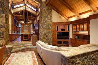 A luxurious open-plan living space in The Treehouse on West Fork features a spacious lounge with a cream sofa, a built-in entertainment center, a full bar, and a gourmet kitchen, all accented by rustic stone and wood elements.