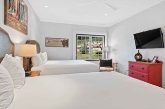 A double queen bedroom in The Charter B1305 featuring two queen-sized beds with white bedding, a wall-mounted TV, a red dresser, and a window overlooking a green outdoor area.