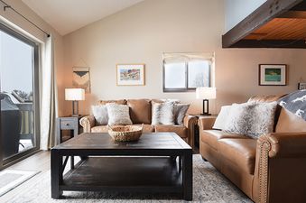 A spacious and inviting living room at Meadow Ridge 19 Unit 1, featuring comfortable leather couches, a dark wood coffee table, and an abundance of natural light streaming in from the large glass door leading to a balcony, showcasing a snowy outdoor scene