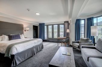A spacious king corner room at The Pines Lodge with a king bed, living area with sofa and armchair, and a desk, all with mountain views through large windows.
