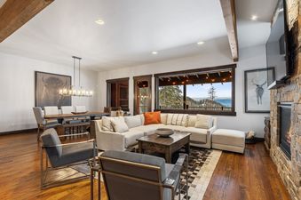A spacious and elegantly furnished living and dining area in Inspiration Ski Haus, featuring a large sectional sofa, a stone fireplace, and a dining table, all set against a backdrop of panoramic snow-covered mountain views through a grand window.