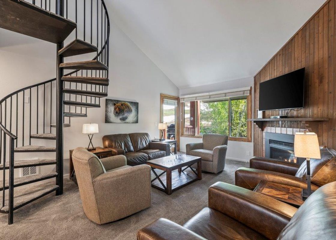 A spacious and inviting living room at Snow Flower 79 Park City, featuring comfortable leather seating, a spiral staircase, and a large window overlooking a verdant outdoor landscape.